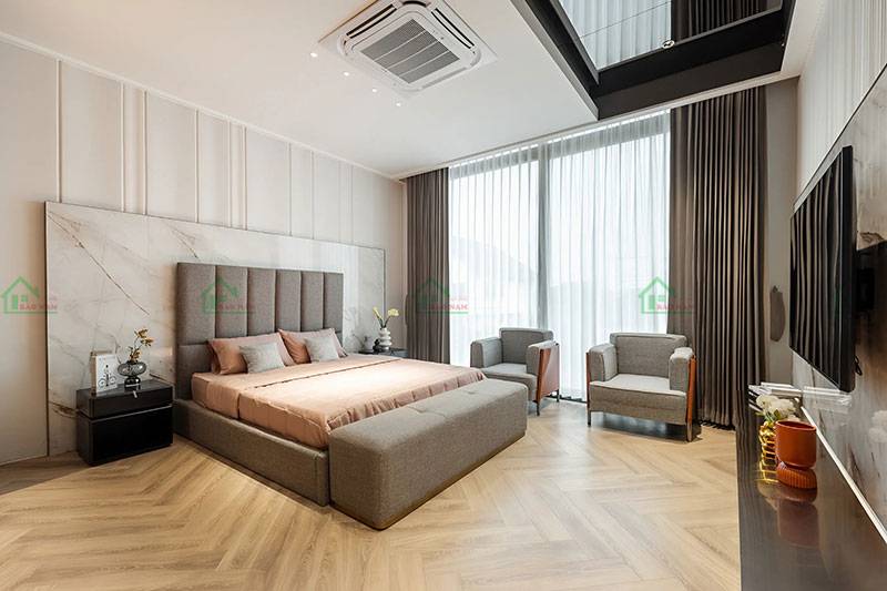 Phòng ngủ sang trọng với thiết kế tinh tế, giường ngủ màu be, đầu giường ốp đá cẩm thạch, và sàn nhà lát gỗ kiểu xương cá độc đáo