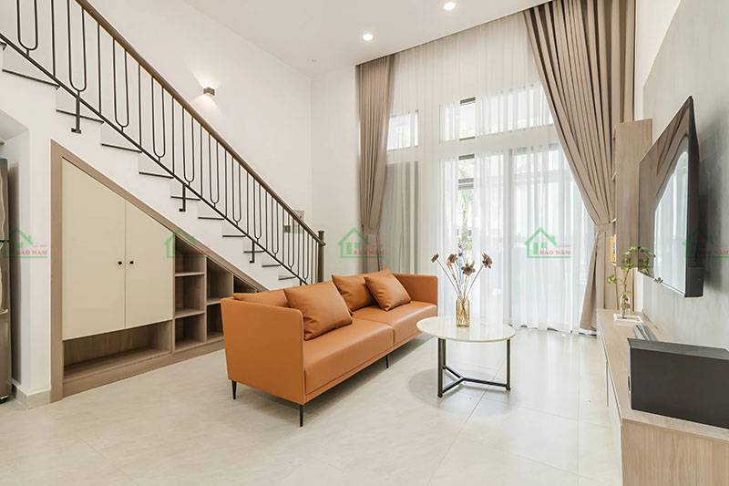 Phòng khách nhà phố thiết kế mở, với sofa dài màu cam đất, kệ tivi âm tường, tường trang trí kết hợp màu xám và cam đất.