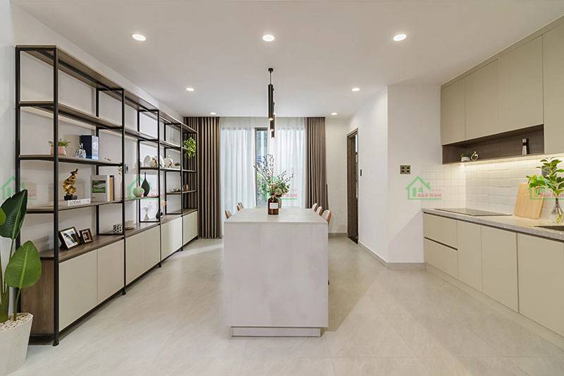 Bố cục mở kết hợp bếp và phòng ăn, với kệ sách, bàn ăn, và tủ bếp màu kem tạo nên không gian liền mạch