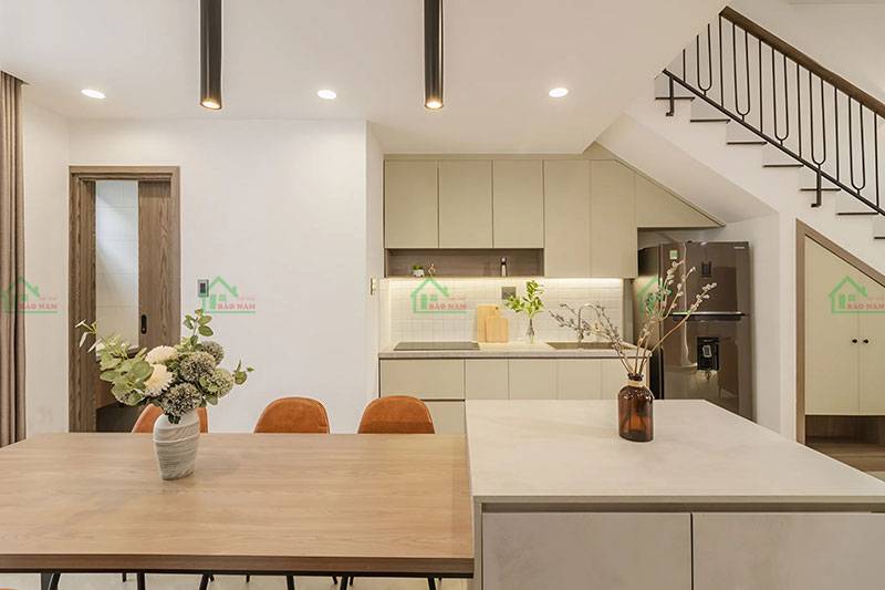 Thiết kế nhà bếp và phòng ăn hiện đại, tối giản, với bàn ăn gỗ, tủ bếp màu kem, và đèn thả tạo điểm nhấn
